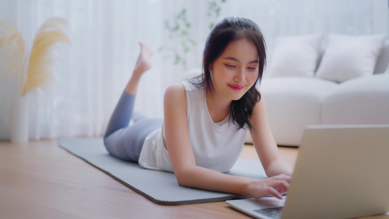 年轻的亚洲女性躺在地板上的瑜伽垫上，坐在笔记本电脑前，在家训练。美丽的微笑的女人在做伸展运动，同时在锻炼前与教练在线交流视频素材画面2