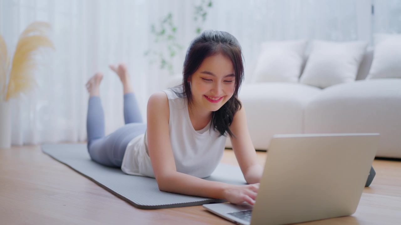 年轻的亚洲女性躺在地板上的瑜伽垫上，坐在笔记本电脑前，在家训练。美丽的微笑的女人在做伸展运动，同时在锻炼前与教练在线交流视频素材画面3