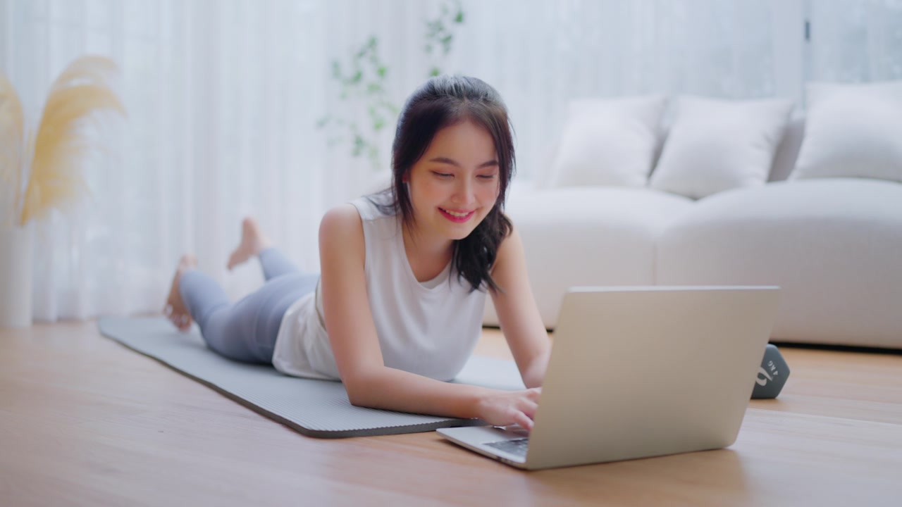 年轻的亚洲女性躺在地板上的瑜伽垫上，坐在笔记本电脑前，在家训练。美丽的微笑的女人在做伸展运动，同时在锻炼前与教练在线交流视频素材画面4