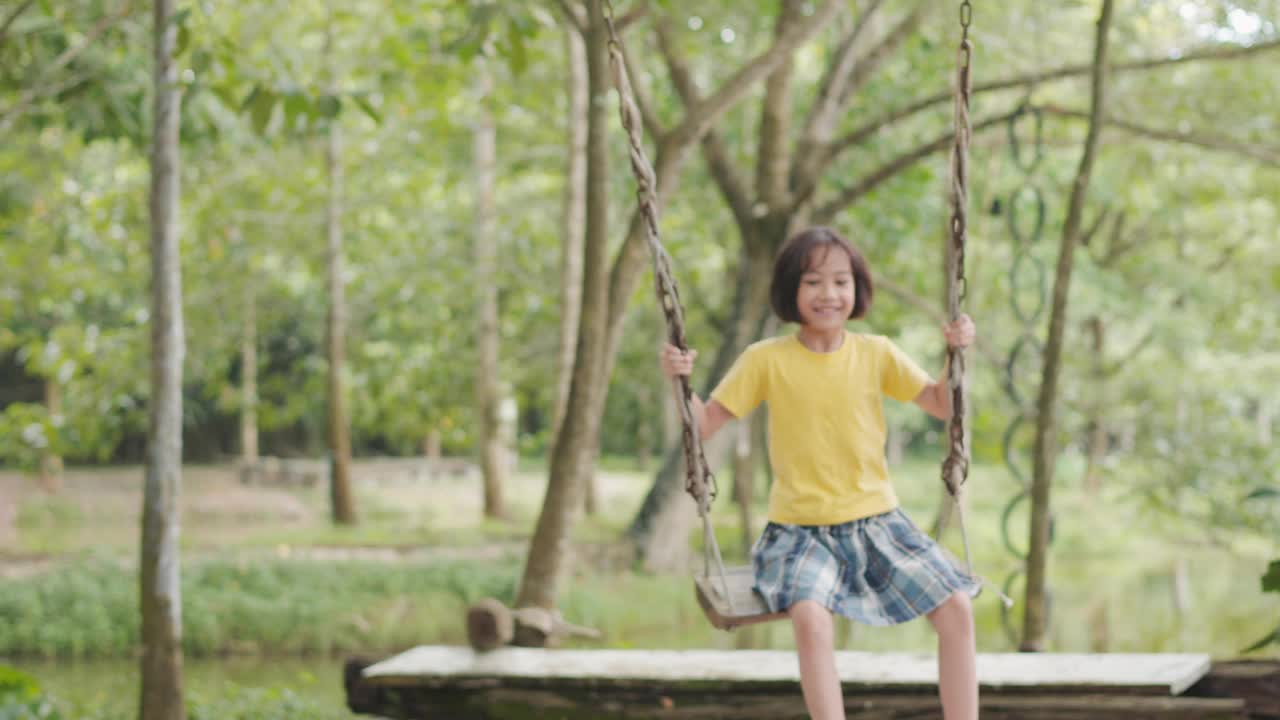 可爱的亚洲小女孩在树木环绕的户外欢快地荡秋千。它表现了童年玩耍、快乐和冒险的本质，突出了自然的无忧无虑的精神和宁静。视频素材