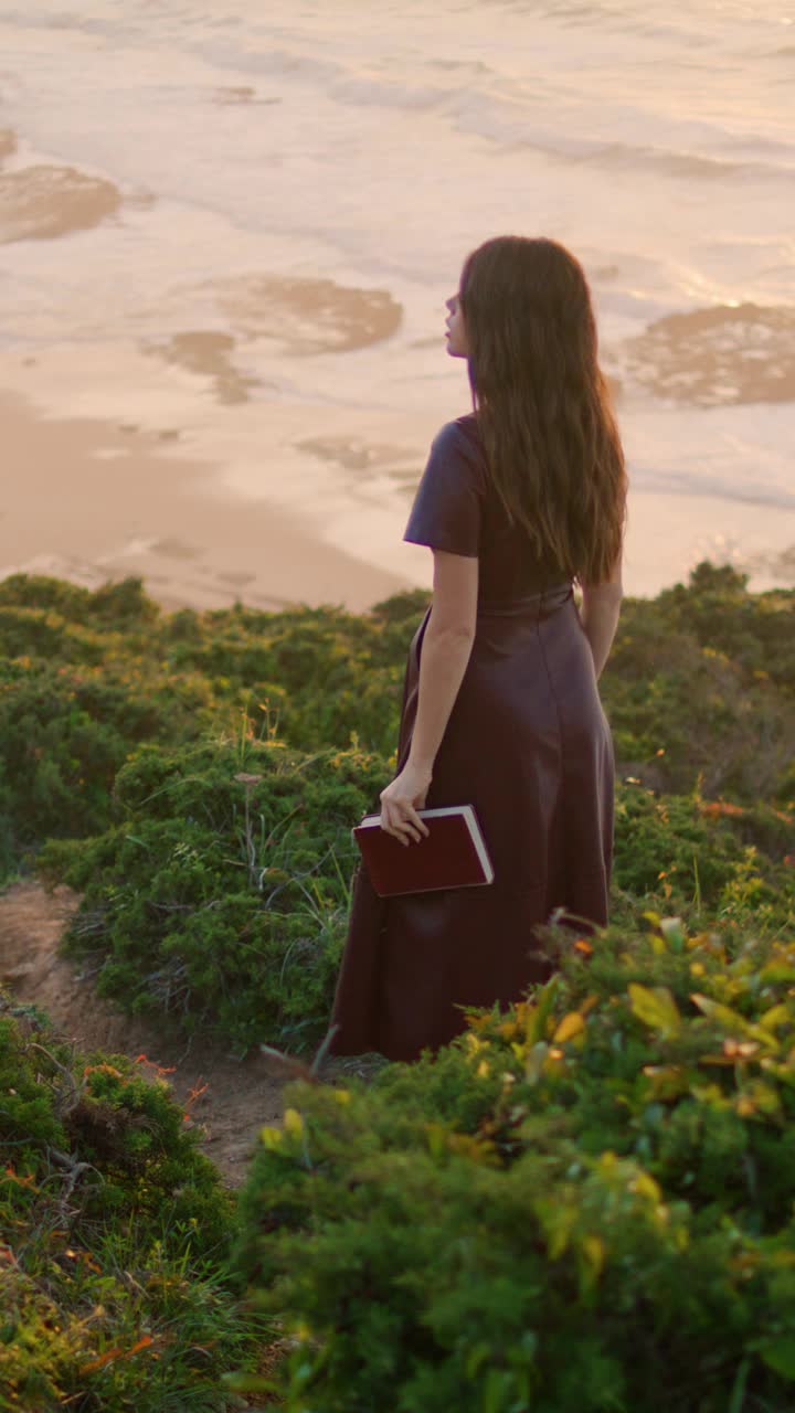 一个不知名的女人直立地拿着书走在山上。女人享受黄昏海岸视频素材