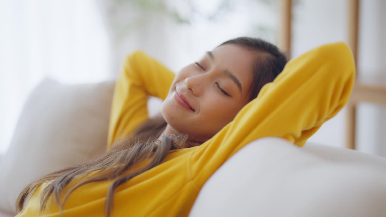 放松的年轻亚洲女人在舒适的沙发上享受休息，平静迷人的女孩在沙发上放松，闭上眼睛呼吸新鲜空气，在家冥想视频素材画面1