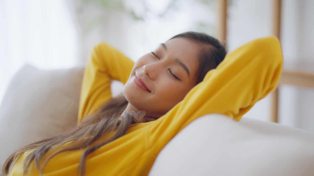 放松的年轻亚洲女人在舒适的沙发上享受休息，平静迷人的女孩在沙发上放松，闭上眼睛呼吸新鲜空气，在家冥想视频素材画面2