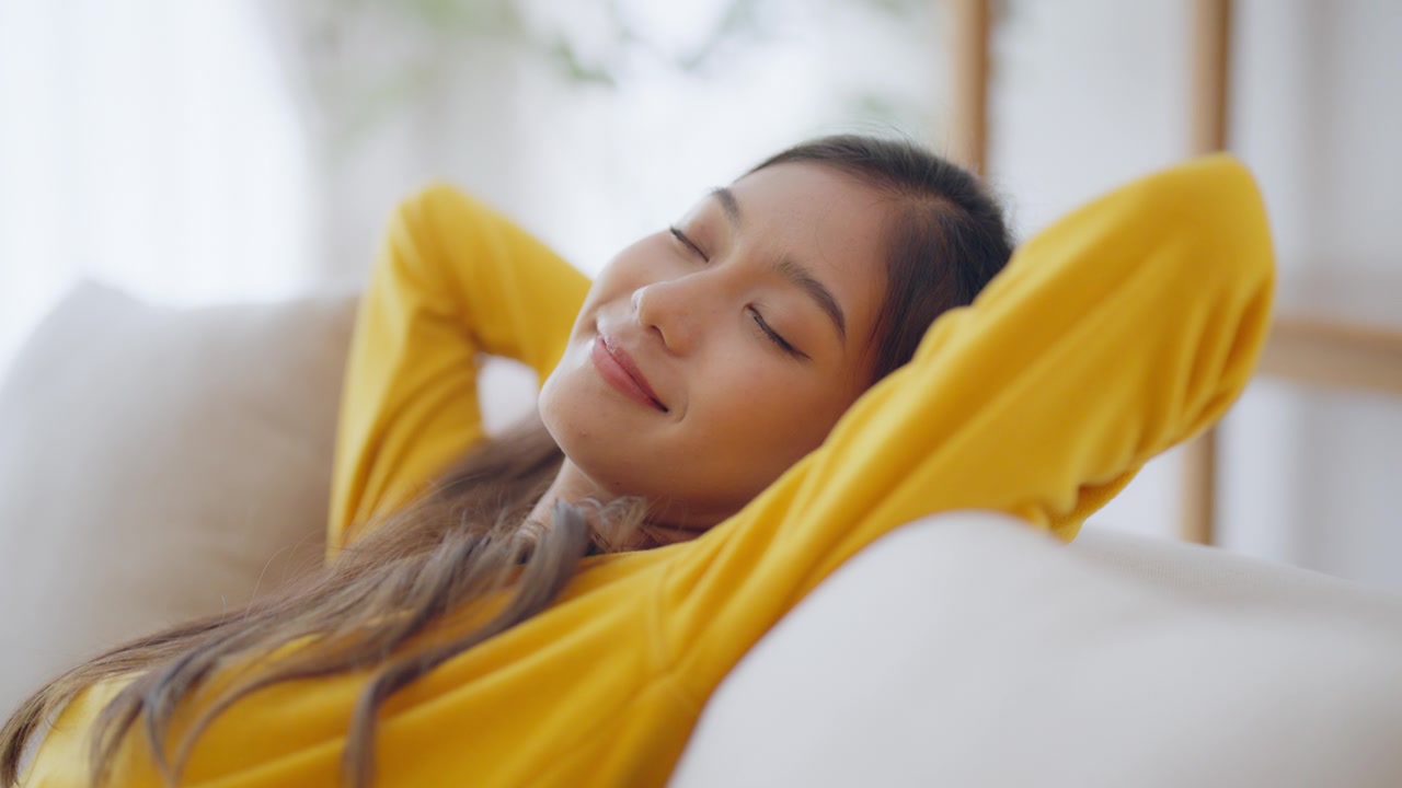 放松的年轻亚洲女人在舒适的沙发上享受休息，平静迷人的女孩在沙发上放松，闭上眼睛呼吸新鲜空气，在家冥想视频素材画面3