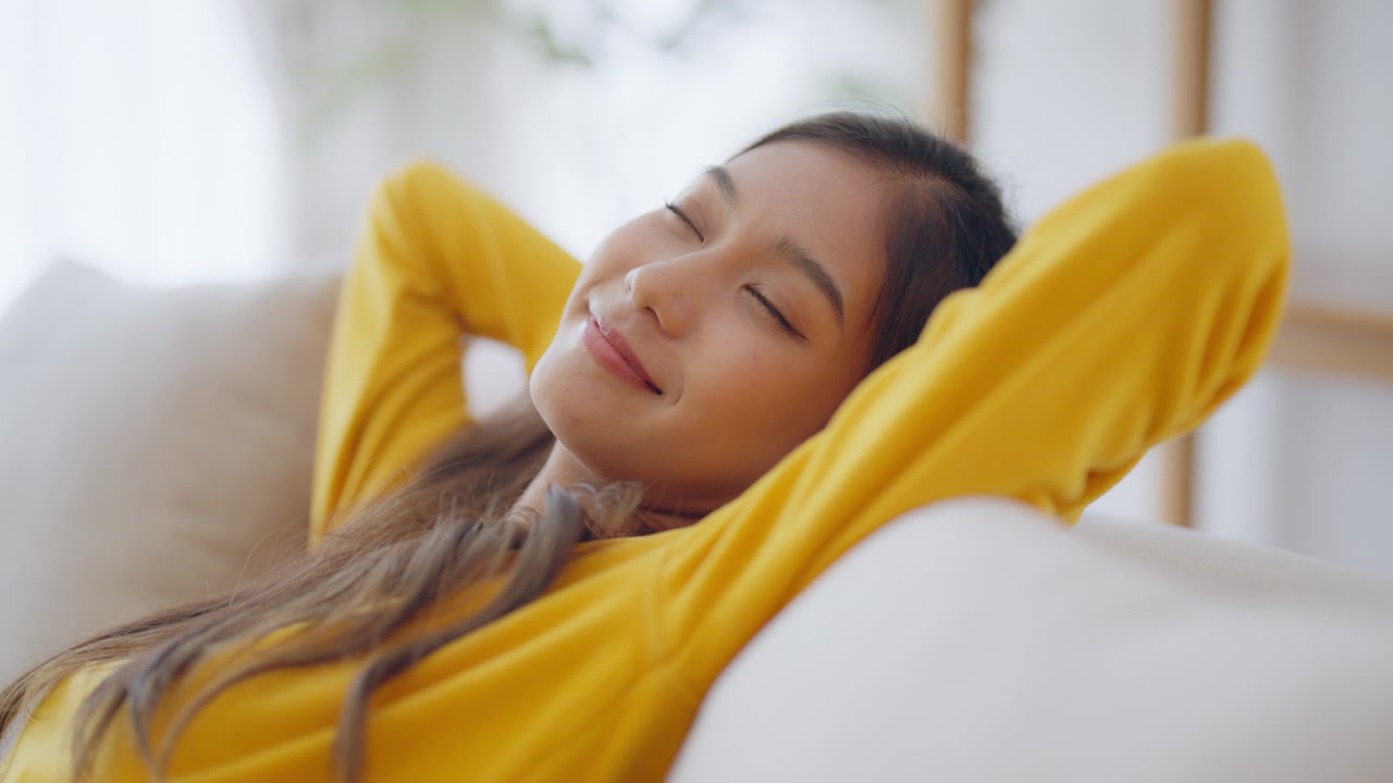放松的年轻亚洲女人在舒适的沙发上享受休息，平静迷人的女孩在沙发上放松，闭上眼睛呼吸新鲜空气，在家冥想视频素材画面4