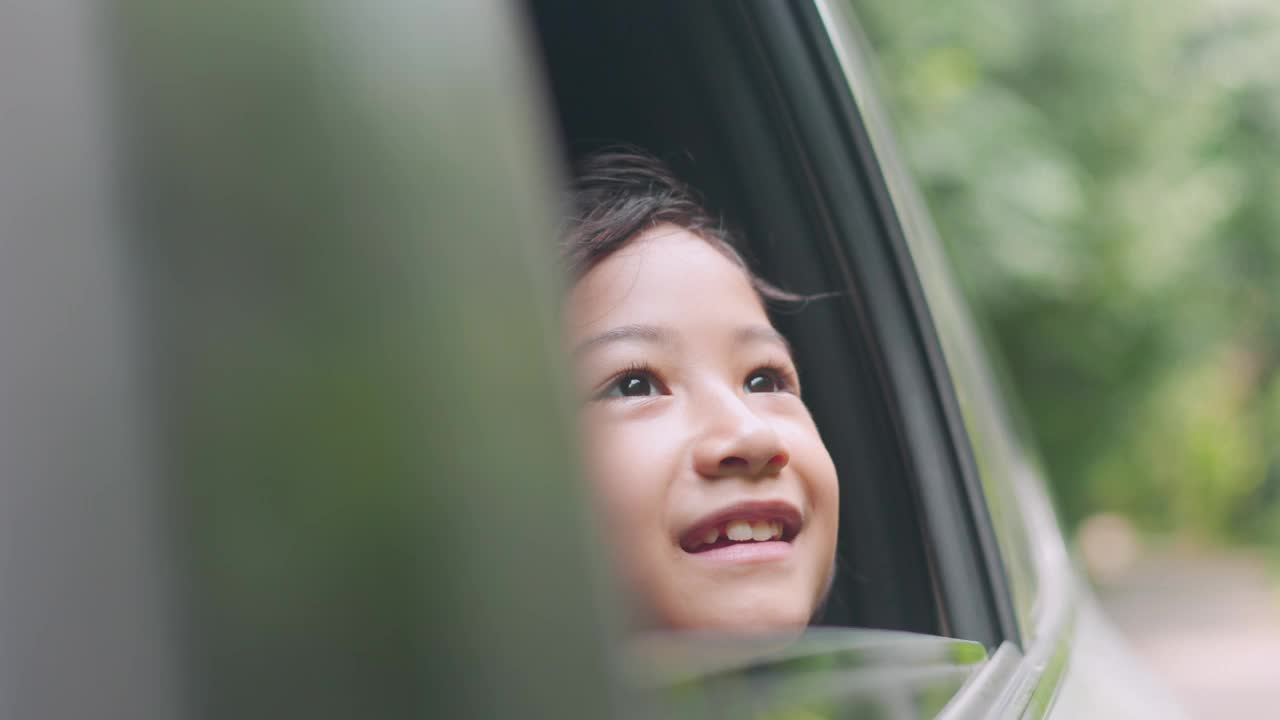 一个可爱的亚洲女孩凝视着车窗外的快乐公路旅行。在郁郁葱葱的绿色背景下，它展示了旅行、家庭、自然和快乐时刻的冒险、好奇心和童年的天真。视频素材