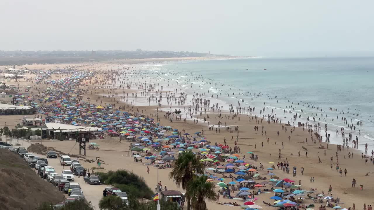 康尼尔德拉弗朗特拉海滩的全景，加的斯，在一个多云的日子里，一群游泳者和五颜六色的遮阳伞覆盖在沙滩上，而许多游客享受水视频下载