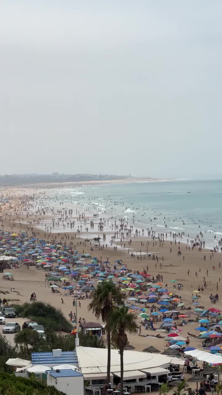 康尼尔德拉弗朗特拉海滩的全景，加的斯，在一个多云的日子里，一群游泳者和五颜六色的遮阳伞覆盖在沙滩上，而许多游客享受水视频下载