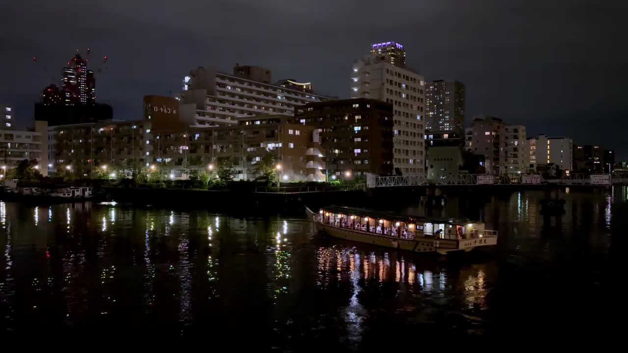 夜晚，一艘传统的船带着灯光在东京的河上巡游，背景是城市建筑视频下载