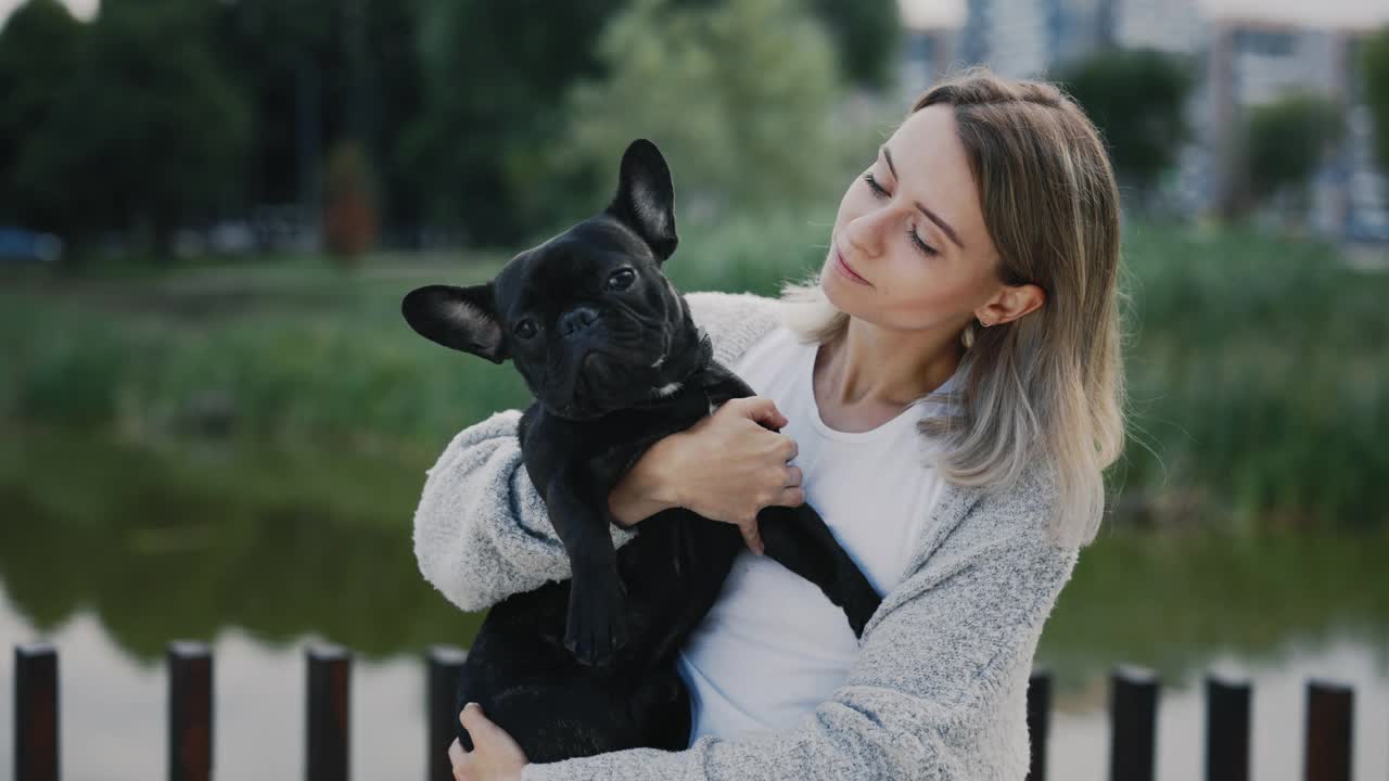 幸福微笑的女人的肖像，抱着她的黑色法国斗牛犬小狗，看着相机在当地的湖泊和公园的背景。人和狗的友谊概念。视频素材