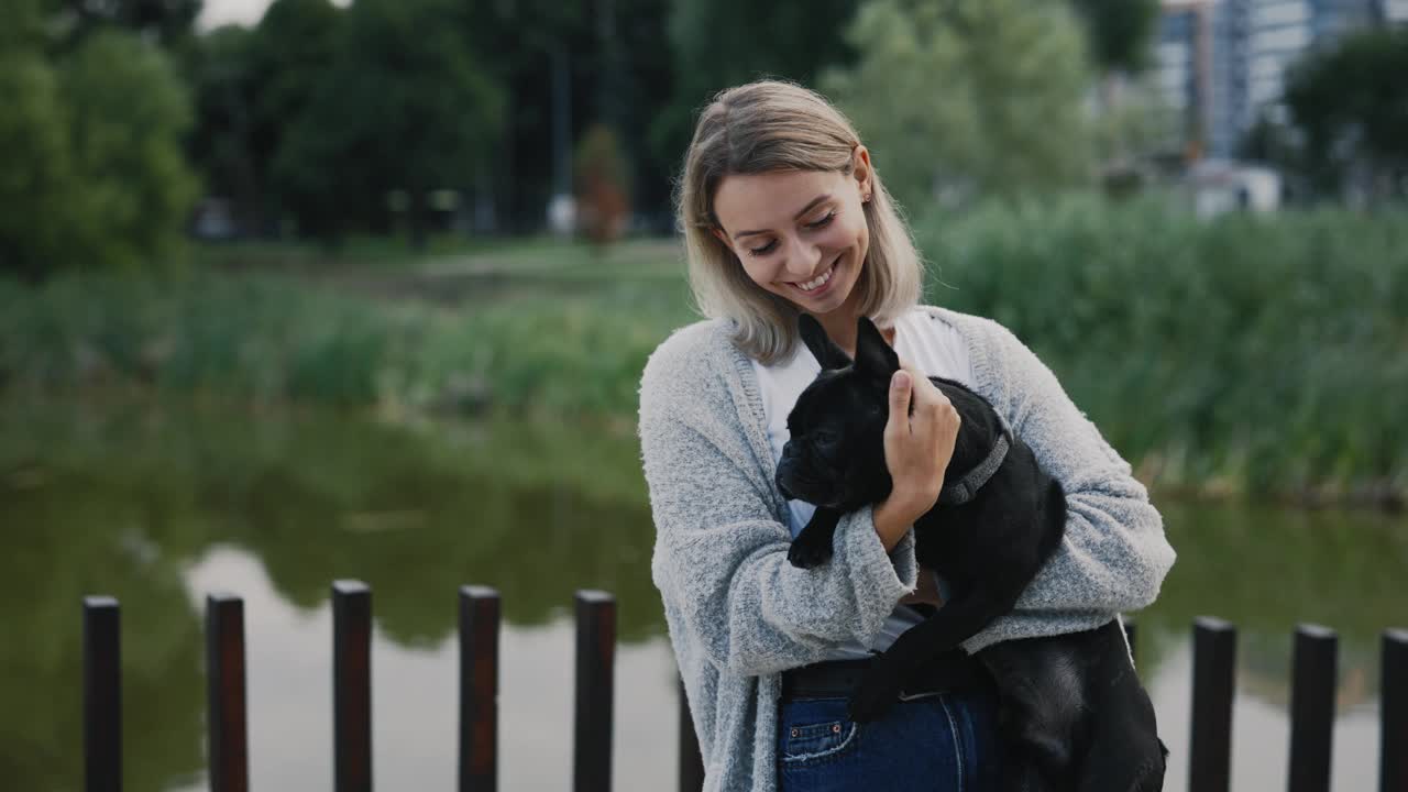 幸福微笑的女人的肖像，抱着她的黑色法国斗牛犬小狗，看着相机在当地的湖泊和公园的背景。人和狗的友谊概念。视频素材