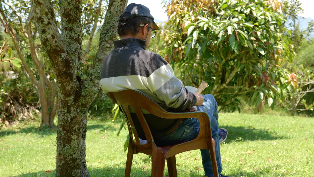 一个老人在阳光明媚的日子里，坐在椅子上，在蓝天下看书，周围是大自然、山脉和森林视频下载