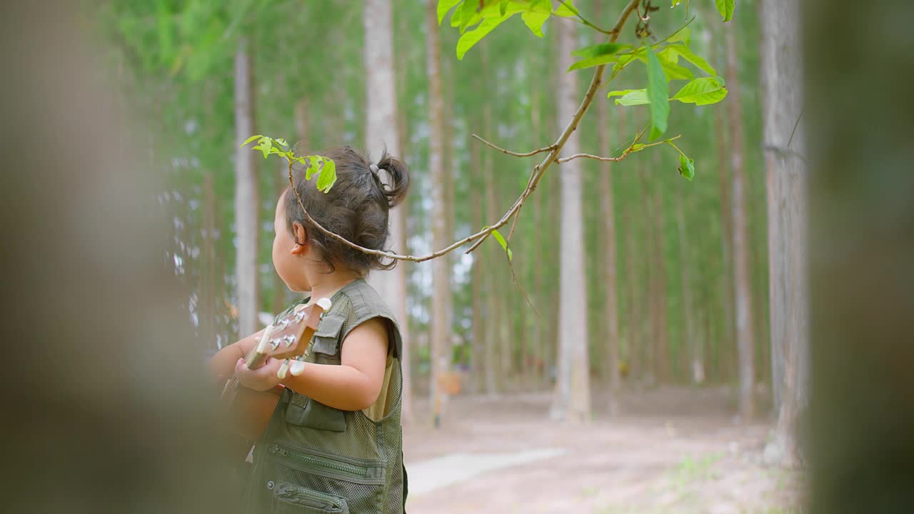 一个快乐的亚洲孩子在宁静的森林中演奏着四弦琴，周围是绿色植物和树木视频素材