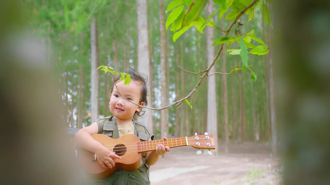 一个快乐的亚洲孩子在宁静的森林中演奏着四弦琴，周围是绿色植物和树木视频素材