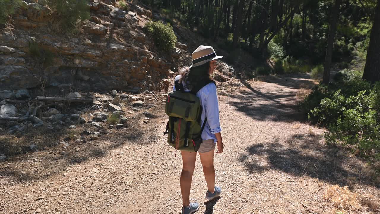 一名女子背着背包，戴着帽子，在森林小径上冒险徒步旅行，享受着阳光明媚的日子和大自然视频素材