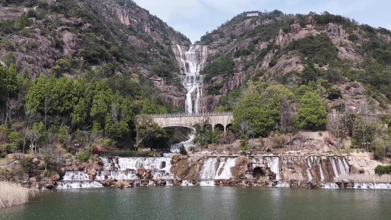 航拍无人机视角：天台山瀑布倾泻而下，越过崎岖的山崖，汇入宁静的湖泊，一座雅致的石拱桥横跨其上，四周环绕着郁郁葱葱的绿叶，天空晴朗视频素材画面1