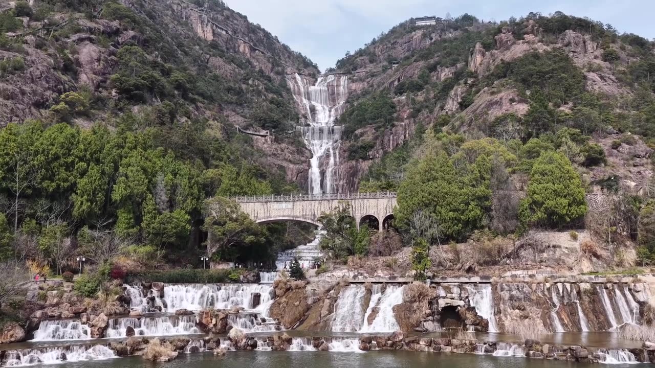 航拍无人机视角：天台山瀑布倾泻而下，越过崎岖的山崖，汇入宁静的湖泊，一座雅致的石拱桥横跨其上，四周环绕着郁郁葱葱的绿叶，天空晴朗视频素材画面2