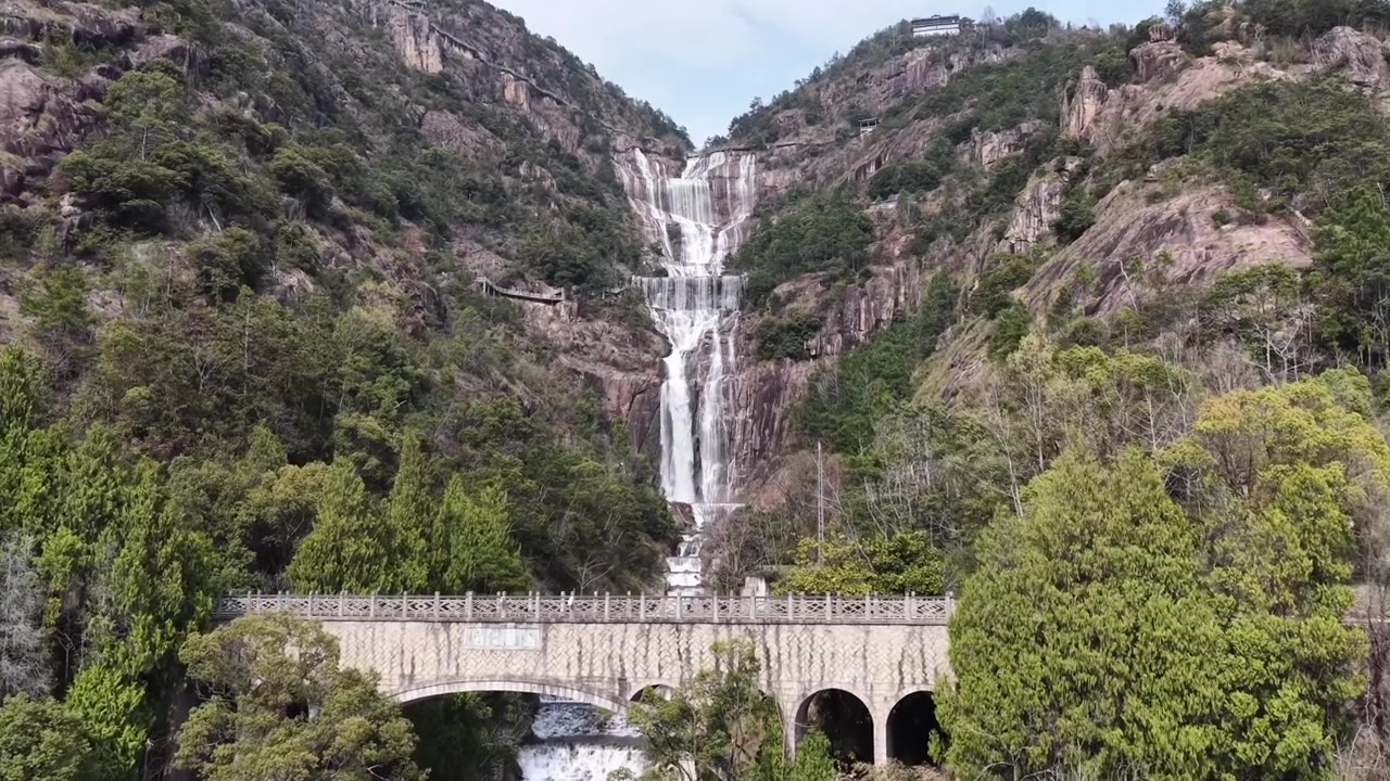 航拍无人机视角：天台山瀑布倾泻而下，越过崎岖的山崖，汇入宁静的湖泊，一座雅致的石拱桥横跨其上，四周环绕着郁郁葱葱的绿叶，天空晴朗视频素材画面4