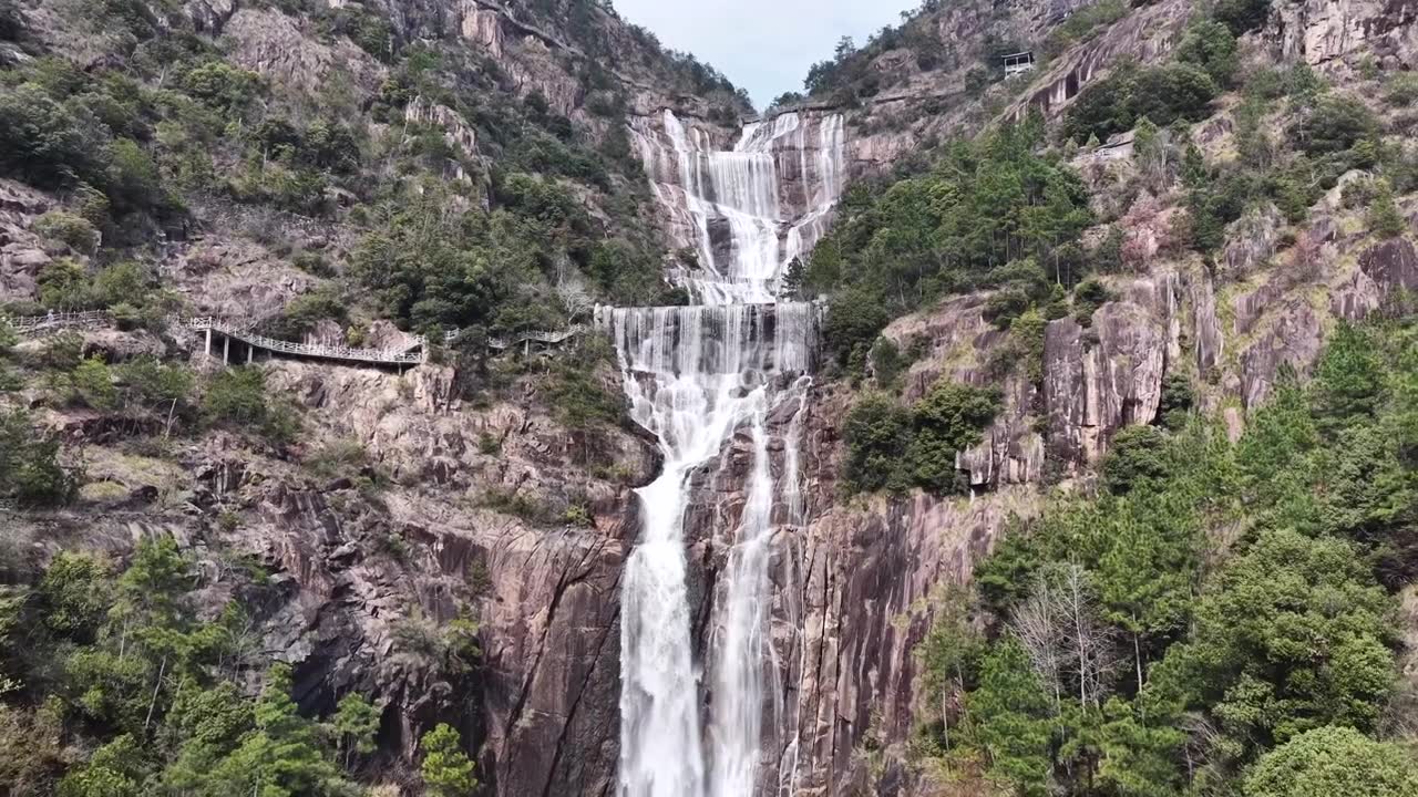 航拍无人机视角，浙江天台山，多层瀑布沿着崎岖的岩石层层叠叠落下，陡峭的山坡，周围是充满活力的绿色植物，蜿蜒的木制步道供徒步旅行者行走。视频素材画面1