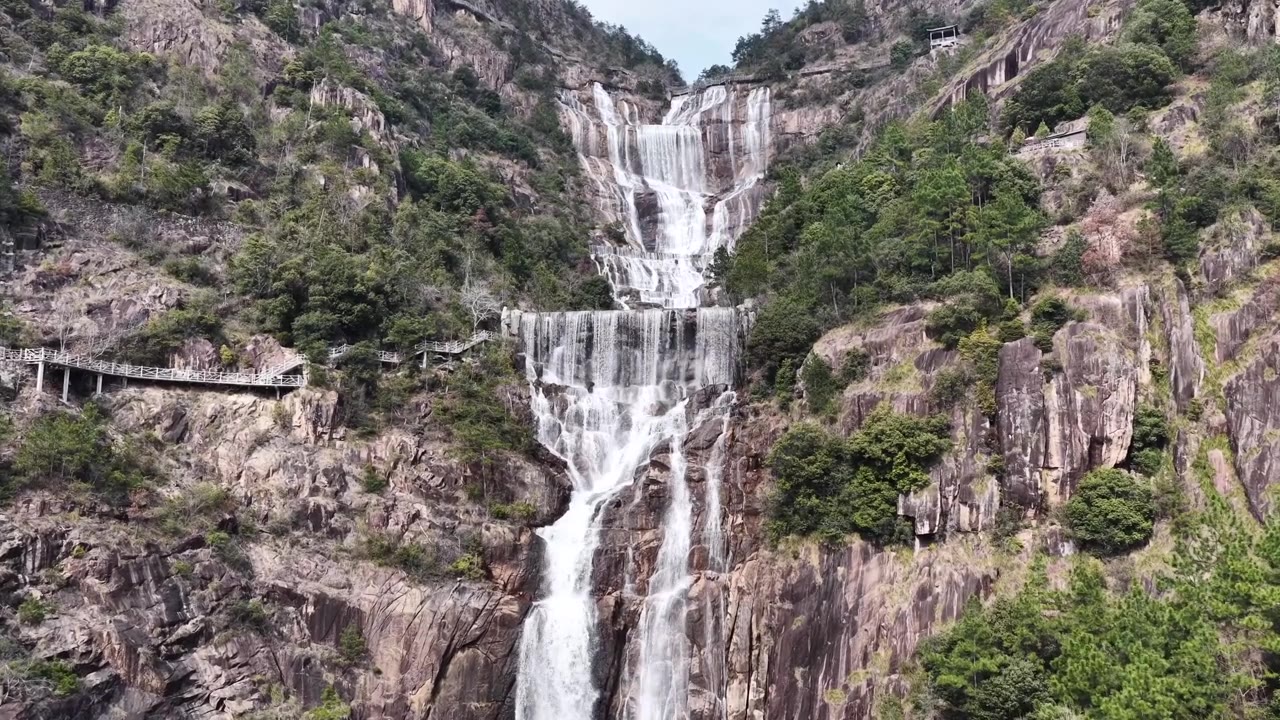 航拍无人机视角，浙江天台山，多层瀑布沿着崎岖的岩石层层叠叠落下，陡峭的山坡，周围是充满活力的绿色植物，蜿蜒的木制步道供徒步旅行者行走。视频素材画面3