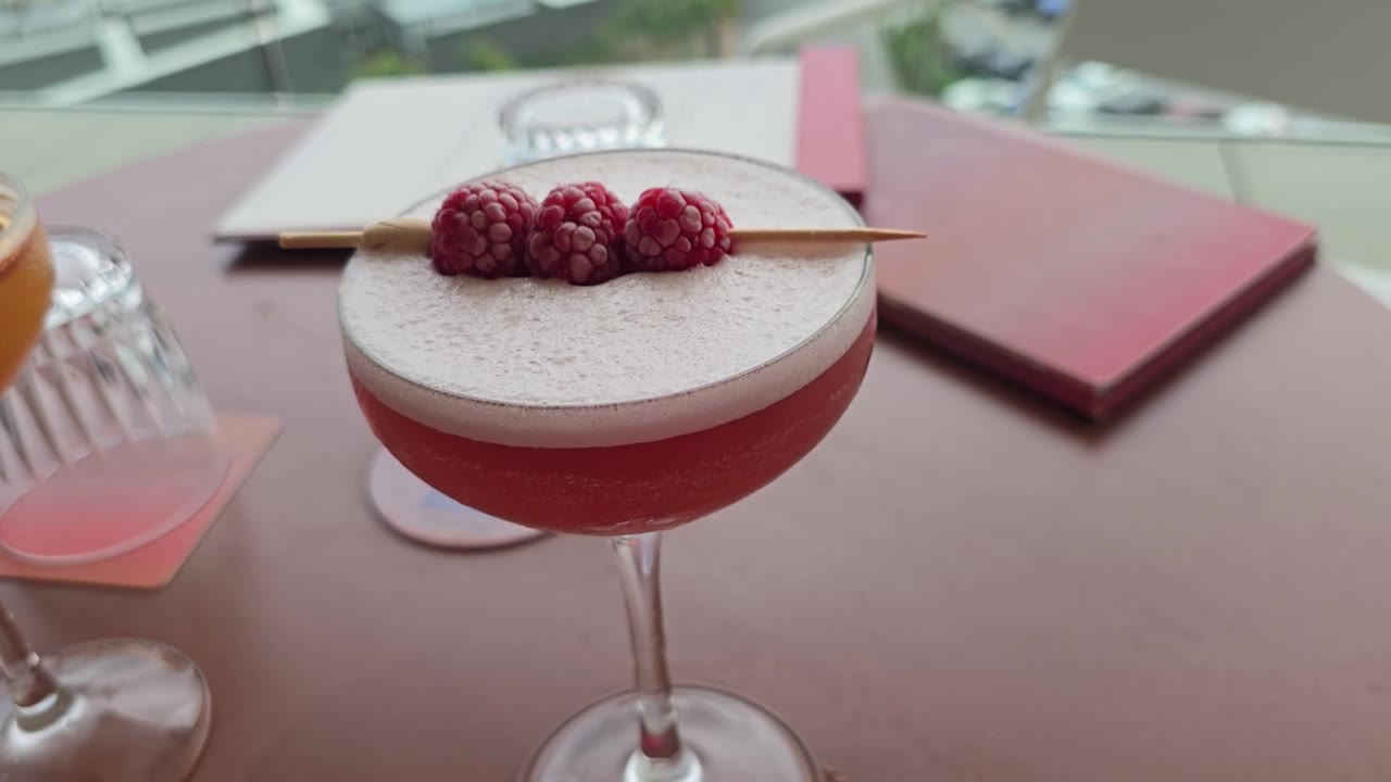 一家屋顶酒吧，桌上放着一杯鸡尾酒，上面插着一串覆盆子，背景是菜单和城市景观。视频素材画面1
