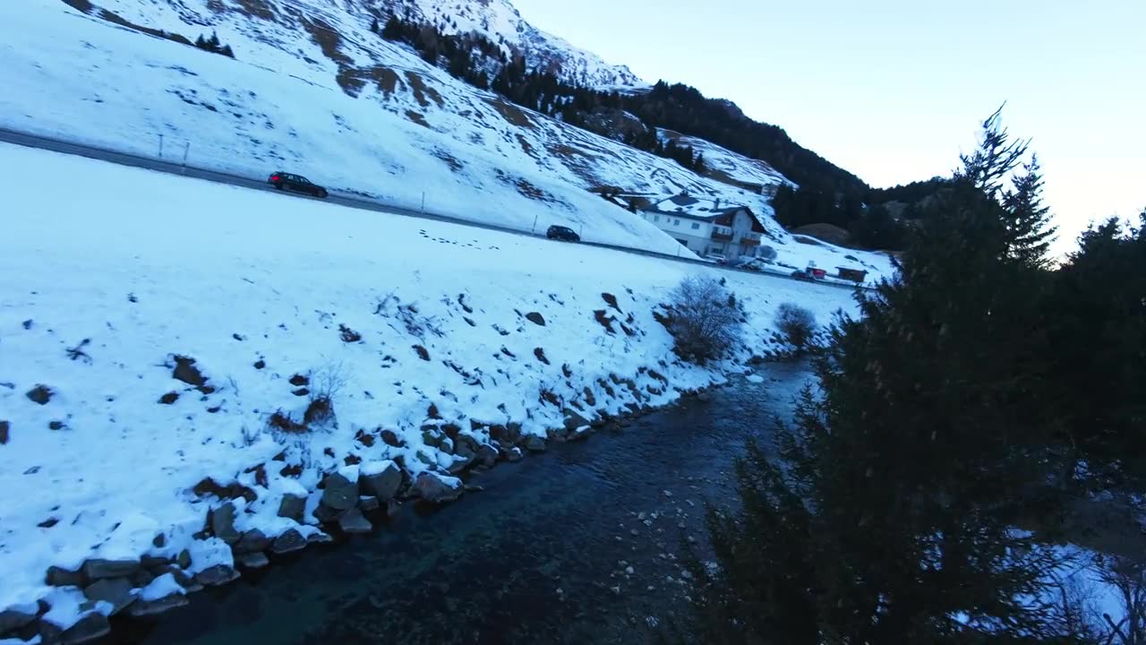 瑞士朱利尔山口，FPV穿越机沿蜿蜒河流向前飞行，穿过白雪皑皑的高山山谷，经过路边斜坡，展现宁静的冬季山景视频素材画面1