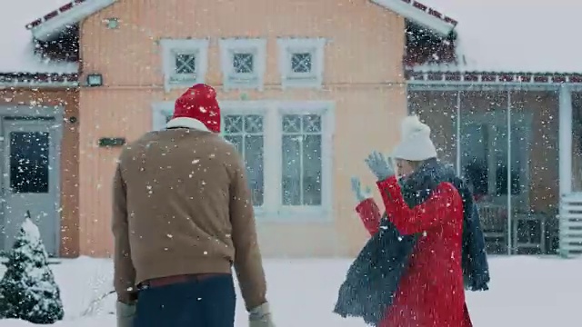 年轻的夫妇顽皮地把雪扔在空中，而冬天下雪继续。快乐的男人和女人在他们田园诗般的房子的院子里玩雪。家庭享受冬天。视频素材