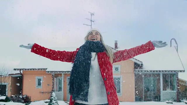 快乐的年轻微笑的女人把雪抛向空中。穿红外套戴围巾的女孩在自家后院享受冬天。视频素材