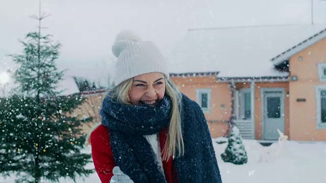 快乐的年轻微笑的女人扔雪球，并被一个击中。穿红外套的女孩和她的伴侣享受冬天。田园小屋后院里的一天。视频素材