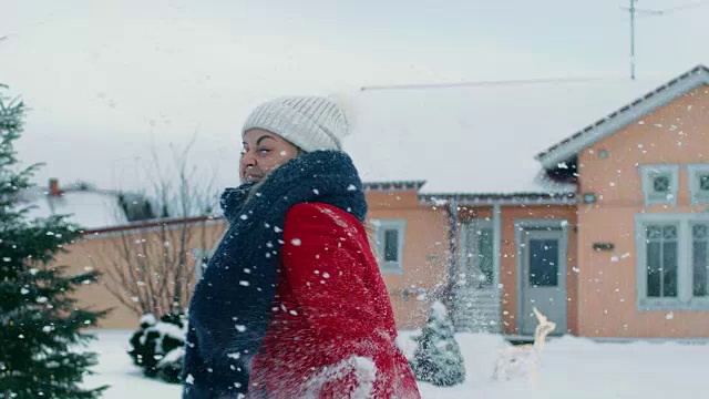 快乐的年轻微笑的女人扔雪球，并被一个击中。穿红外套的女孩和她的伴侣享受冬天。在缓慢的运动。田园小屋后院里的一天。视频素材