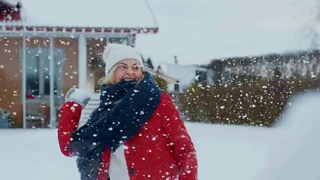 快乐的年轻微笑的女人扔雪球，并被一个击中。穿红外套的女孩和她的伴侣享受冬天。田园小屋后院里的一天。视频素材