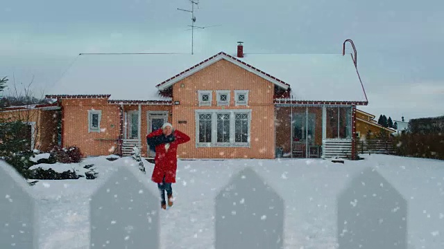 美丽的年轻微笑女人跳跃和旋转下飘落的雪。穿红外套戴围巾的女孩在后院享受冬天。田园诗般的房子站在背景。视频素材