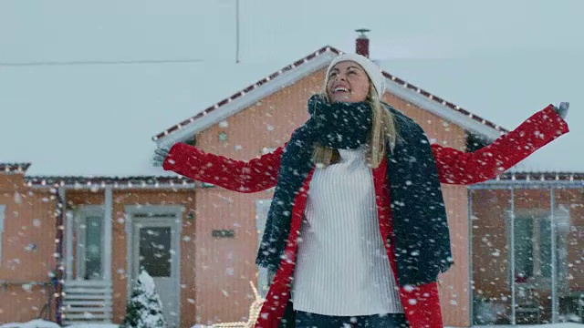 美丽微笑的年轻女子在飘落的雪中旋转。穿红外套戴围巾的女孩在院子里享受冬天。视频素材