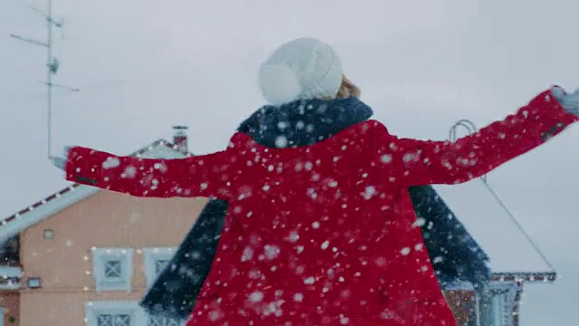 美丽微笑的年轻女子在飘落的雪中旋转。穿红外套戴围巾的女孩在院子里享受冬天。视频素材