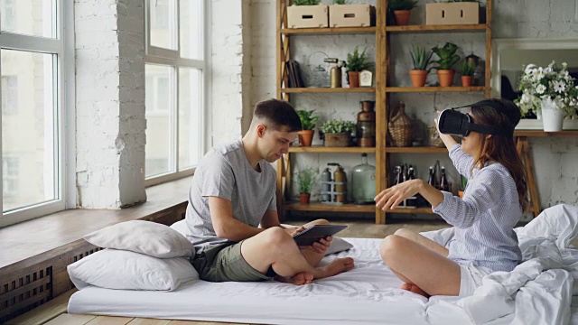 一对相爱的夫妇正在享受增强现实眼镜，女人戴着它们移动手臂和身体，而她的丈夫使用平板触屏看着他的妻子。视频素材画面1